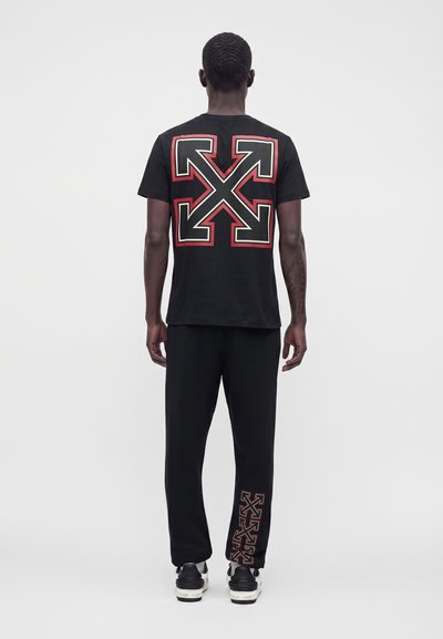 Homme debout de dos, portant un t-shirt noir avec un grand motif flèche rouge et blanc et un pantalon noir avec un motif flèche assorti sur la jambe.