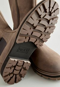 Brown leather boots with a textured synthetic sole; visible tread pattern and zipper detail on the side. Brand name 'NEXT' embossed on the sole.