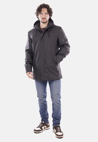 Giacca esterna nera, impermeabile, con cappuccio, dotata di tasche con zip e vestibilità slim, abbinata a jeans blu e sneakers marroni.