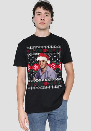 Junger Mann trägt ein schwarzes Urlaubsmotiv-T-Shirt mit Elvis Presley in Weihnachtsmannmütze und Weihnachtsmustern in Rot, Grün und Weiß.