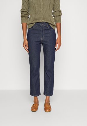 Osoba ubrana w ciemnoniebieskie dżinsy o prostych nogawkach, oliwkowozielony sweter z grubym splotem i brązowe mokasyny stoi na tle jednolitego tła.