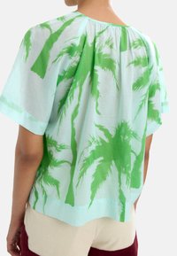 Zielony nadruk palm na jasnoniebieskim materiale. Krótkie rękawy i luźny krój. Teksturowany, zwiewny materiał z casualowym dekoltem.