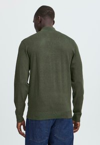 Mørkegrønn strikket genser med lange ermer, rund hals og ribbestrikket detalj ved kantene og mansjettene. Brukt over blå denimbukser.