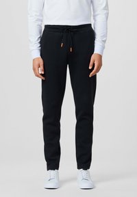 Zwarte joggingsbroek met een trekkoord in de taille, voorzien van oranje uiteinden, gecombineerd met witte sneakers en een lange mouwen witte shirt.