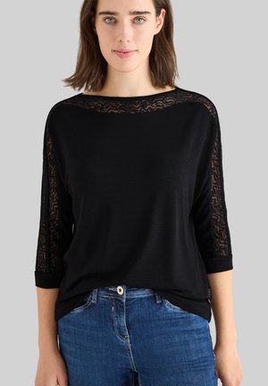 Schwarzes Oberteil mit Drei-Viertel-Ärmeln, das an den Schultern mit einem transparenten gemusterten Netzdesign versehen ist. Kombiniert mit blauen Jeans.