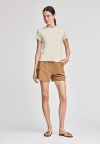 T-shirt beige a maniche corte con un piccolo logo, abbinata a pantaloni cargo color cammello con tasche laterali e ciabatte color marrone chiaro.