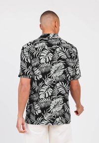 Camisa negra de manga corta con un patrón de hojas tropicales blancas. Tejido ligero y diseño de cuello clásico visible desde la parte de atrás.