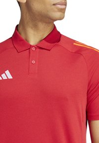 Rode polo shirt met een textuurstof, een kraag met twee knopen en drie witte strepen op de linker schouder.
