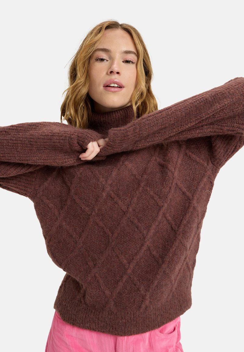 Brauner gestrickter Pullover mit einem hohen Kragen, der ein strukturiertes Rautenmuster und oversized Ärmel aufweist. Getragen über rosa Hosen.