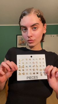 Sada 18 náušnic vystavených na bílém kartonu označeném "PIECES." Zlaté a stříbrné odstíny, různé designy včetně hvězdiček, květin a pecek.