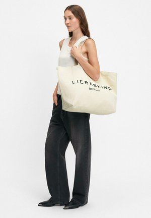 Frau trägt ärmelloses weißes Oberteil und schwarze Jeans mit weitem Bein, hält große beige Tragetasche mit dem Schriftzug "LIEBESKIND BERLIN".