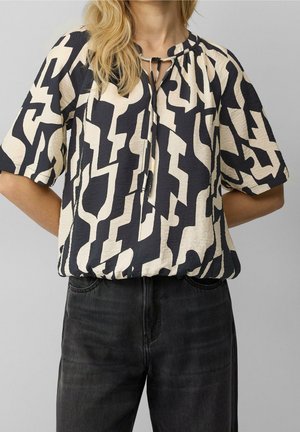 Vrouw draagt een blouse met abstracte print in zwart en beige, korte mouwen en een strikdetail bij de halslijn, gecombineerd met zwarte jeans.