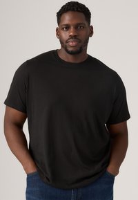 T-shirt noir à manches courtes en tissu doux avec un col rond classique. Porté de manière décontractée avec les mains dans les poches, associé à un jean bleu.