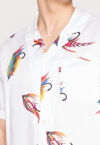 Koszula na krótki rękaw z białego materiału z wielokolorowymi wzorami piór. Posiada kieszeń na piersi z przodu oraz perłowe guziki.