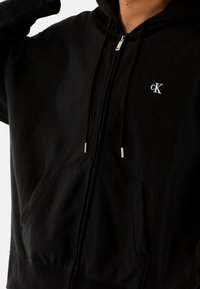 Felpa nera con zip realizzata in tessuto morbido, dotata di tasca a marsupio, cappuccio con coulisse e un piccolo logo bianco "cK" sul petto.