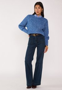 Maglione blu in maglia grossa su una camicia a righe con colletto, abbinato a jeans a gamba larga di un blu scuro e una cintura marrone con grandi dettagli dorati.