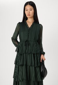 Grön, lager-på-lager, rufsig klänning i transparent material, med v-ringning, långa ärmar och knytning vid midjan. En knuten svart clutch hålls i handen.