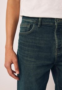 Mørkegrønne denimjeans med et klassisk fem-lomme-design, der har lys syning langs sømmene og en glat, struktureret overflade.