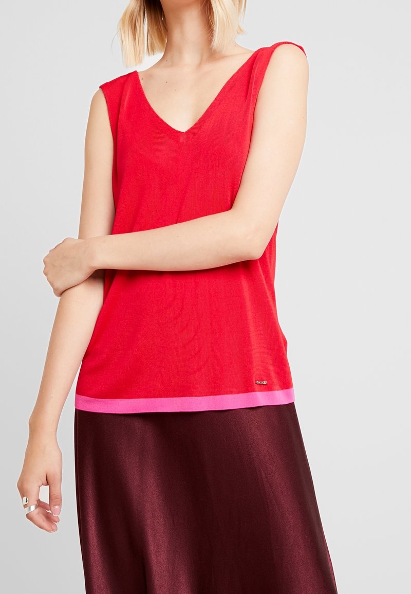 Femme portant un haut rouge sans manches avec un ourlet rose et une jupe bordeaux foncé, debout avec un bras croisé sur l'autre.