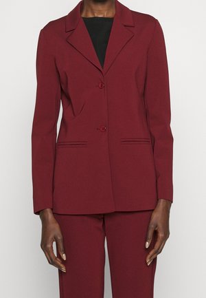 Kobieta ubrana w bordo dopasowaną marynarkę na dwa guziki i dopasowane spodnie, stojąca na tle jednolitego jasnego tła.