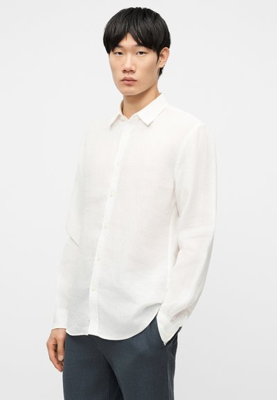 Νεαρός άνδρας με μαύρα μαλλιά που φοράει λευκό πουκάμισο με μακριά μανίκια και σκουρόχρωμο παντελόνι, πόζαροντας με το ένα χέρι στην τσέπη.