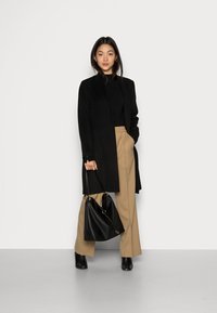 Schwarzer langer Mantel über einem schwarzen Oberteil, beige Hose und schwarze Stiefeletten. Halten einer schwarzen Tasche. Schlichtes Design, glatte Textur.