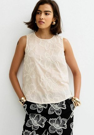 Giovane donna che indossa un top beige senza maniche con ricami floreali e pantaloni neri con motivi floreali bianchi, accessoriata con braccialetti e orecchini dorati.