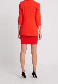 Červený saka na míru s tříčtvrtečními rukávy spárovaný se střiženou červenou sukní. Černé vysoké podpatky dotvářejí outfit. Hladká textura.
