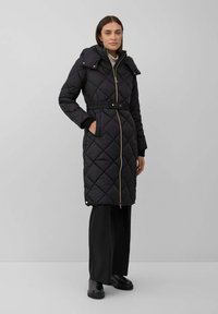 Femme portant un long manteau matelassé noir avec une ceinture, une fermeture éclair et des poches, associé à un pantalon noir et des bottes noires, se tenant contre un mur gris.