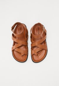 Sandales en cuir marron avec des lanières entrecroisées, design ouvert sur le devant et semelle plate noire. Équipées d'une bride de cheville ajustable avec boucle.
