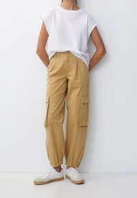 Beżowe spodnie cargo z elastycznymi mankietami, wieloma bocznymi kieszeniami i luźnym fasonem, w połączeniu z luźną białą koszulą i białymi trampkami.