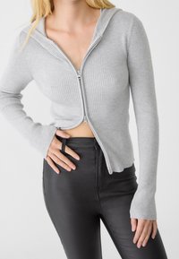 Hoodie gris à fermeture éclair avec un design côtelé ajusté, manches longues et capuche. Associé à un pantalon noir taille haute, affichant un style court.
