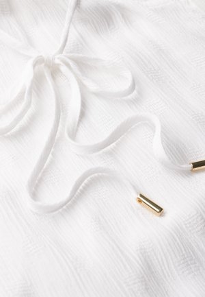 Tissu texturé blanc avec un cordon de serrage noué et des accents métalliques dorés. Le tissu présente un motif subtil, ajoutant de la dimension au design.