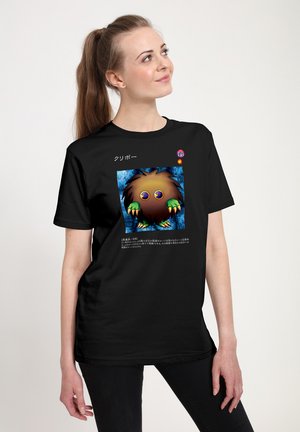 Jeune femme portant un t-shirt noir avec un personnage de dessin animé coloré avec des griffes et de grands yeux, texte japonais au-dessus et en dessous du personnage.