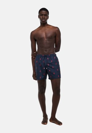 Jeune homme debout pieds nus, portant un short de bain bleu marine avec des motifs de homards rouges, sur un fond blanc uni.