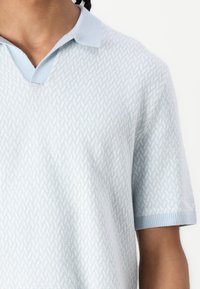 Ανοιχτό μπλε πόλο με υφαντή διαγώνια υφή, κοντά μανίκια και κλασικό γιακά. Το ύφασμα φαίνεται μαλακό και ελαφρύ.