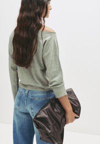 Grauer, schulterfreier Pullover im relaxed Fit, kombiniert mit hochtaillierten blauen Jeans. Hält eine dunkelbraune gefaltete Ledertasche.