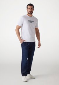 White cotton t-shirt with "GUESS EST. 1981" printed in dark green, paired with dark blue denim trousers and white sneakers. Simple design.