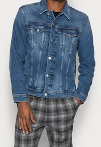 Chaqueta de mezclilla en azul medio con botones plateados, dos bolsillos en el pecho y detalles de costura clásicos. Lleva puesta sobre pantalones a cuadros grises.