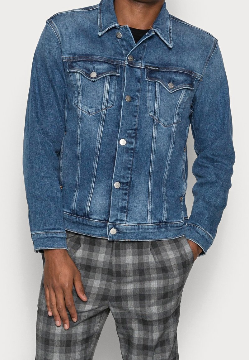 Chaqueta de mezclilla en azul medio con botones plateados, dos bolsillos en el pecho y detalles de costura clásicos. Lleva puesta sobre pantalones a cuadros grises.