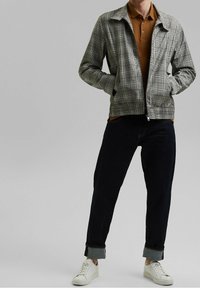 Veste zippée en plaid gris avec des poches latérales, portée sur un polo marron et un jean sombre, complétée par des baskets blanches.