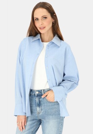 Kobieta ubrana w jasnoniebieską koszulę zapinaną na guziki na biały top oraz jasne dżinsy, pozująca z jedną ręką w kieszeni na białym tle.