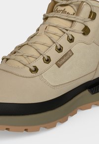 Μπεζ ορειβατικό μποτάκι Timberland με μεταλλικά περντήματα, μπεζ κορδόνια, επενδεδυμένη γλώσσα και ανθεκτική μαύρη και καφέ σόλα.