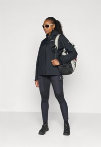 Black waterproof jacket with high collar, paired with dark leggings featuring patch pockets. Both items are designed for outdoor activity. Black backpack.