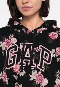 Czarna bluza z kapturem z kwiatowym wzorem w różowym i kremowym kolorze. Posiada duże logo "GAP" w czerni, obszyte różową nicią. Miękka w dotyku tkanina.