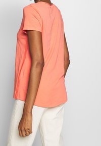 Korálové tričko s krátkým rukávem vyrobené z měkké bavlny, s volným střihem a kulatým výstřihem, spárované s světlými kalhotami.