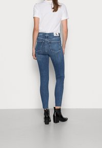Jeans ajustados de color azul con cintura alta, que cuentan con cinco bolsillos y una etiqueta con el logo de CK. El modelo lleva botas de tacón negras hasta el tobillo.