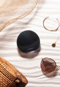 Svart silikon ansiktsrenseenhet med teksturert overflate, sentrert på en lys, bølgende bakgrunn, ledsaget av solbriller og tilbehør.