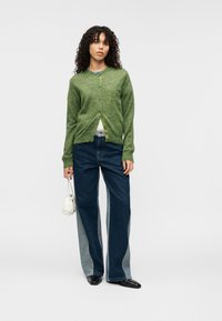 Πράσινη πλεκτή ζακέτα με μπροστινά κουμπιά, σε συνδυασμό με τζιν παντελόνι με φαρδιά μπατζάκια που διαθέτει ανοιχτό και σκούρο τζιν ύφασμα. Κρατάει μια μικρή λευκή τσάντα.