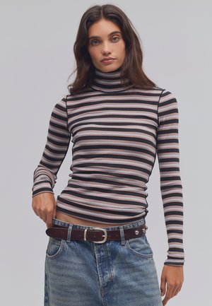 Femme portant un haut à col roulé rayé à manches longues et un jean bleu avec une ceinture marron, posant avec une main près de sa taille.